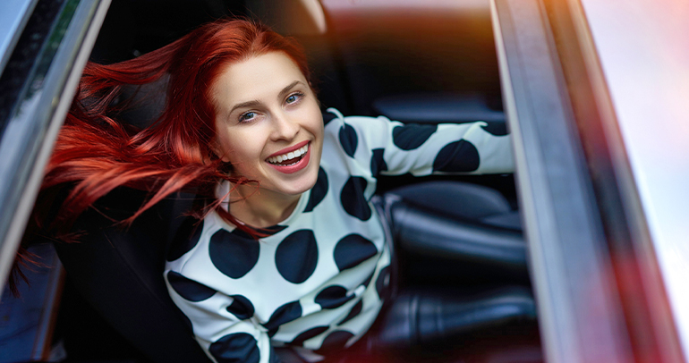 A vibrant, smiling woman looks up through the sunroof of her battery electric vehicle