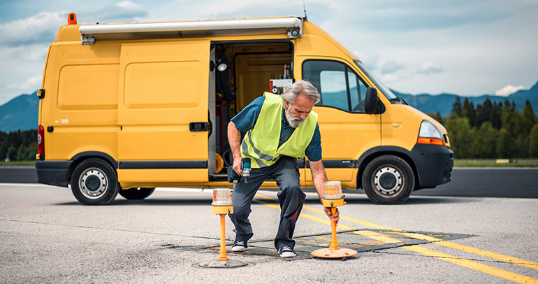 A mature, male airport mechanic is repairing an elevated light using equipment from his yellow box van that is parked behind him