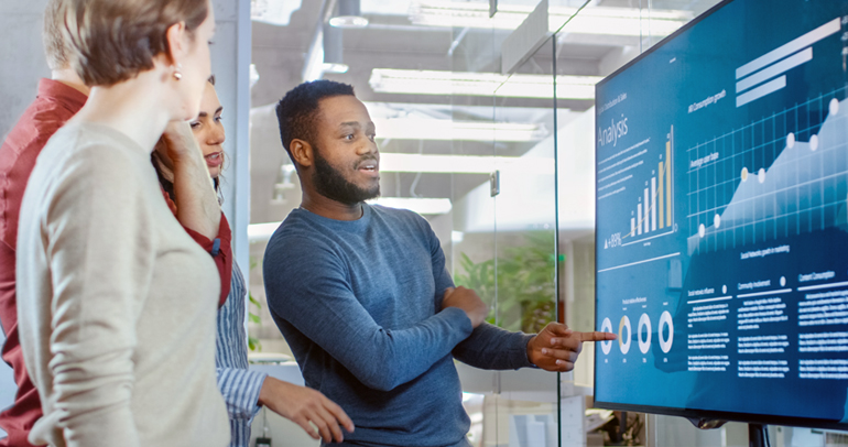 Image of four fleet decision-makers stand around a large monitor considering and discussing data from connected fleet services that are displayed in various charts