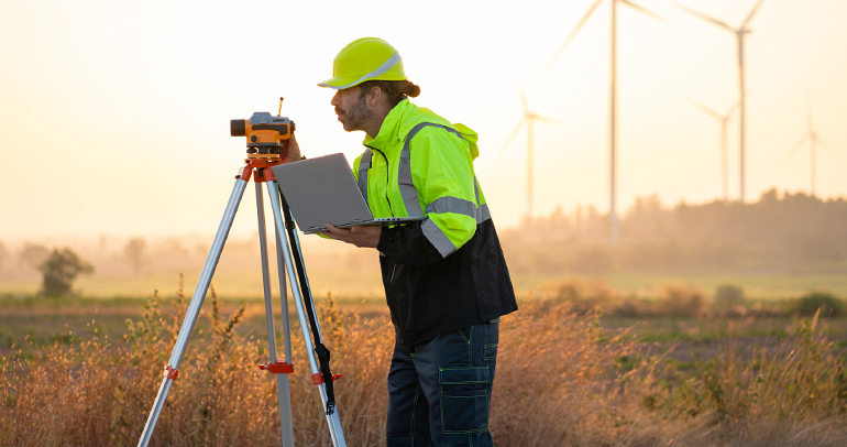 Utility survey worker with laptop and survey stand taking measurements in a wind turbine field