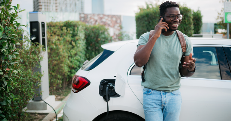 A happy young man leaning on his battery electric vehicle as it charges, talking on his phone with a smile